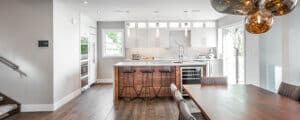 Modern kitchen with white cabinets, a marble backsplash, wooden island with barstools, pendant lights, and a dining table with gray chairs; large windows provide natural light.