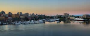 Panoramic view of a marina with boats docked, city buildings in the background, and calm water reflecting the sunrise or sunset sky.