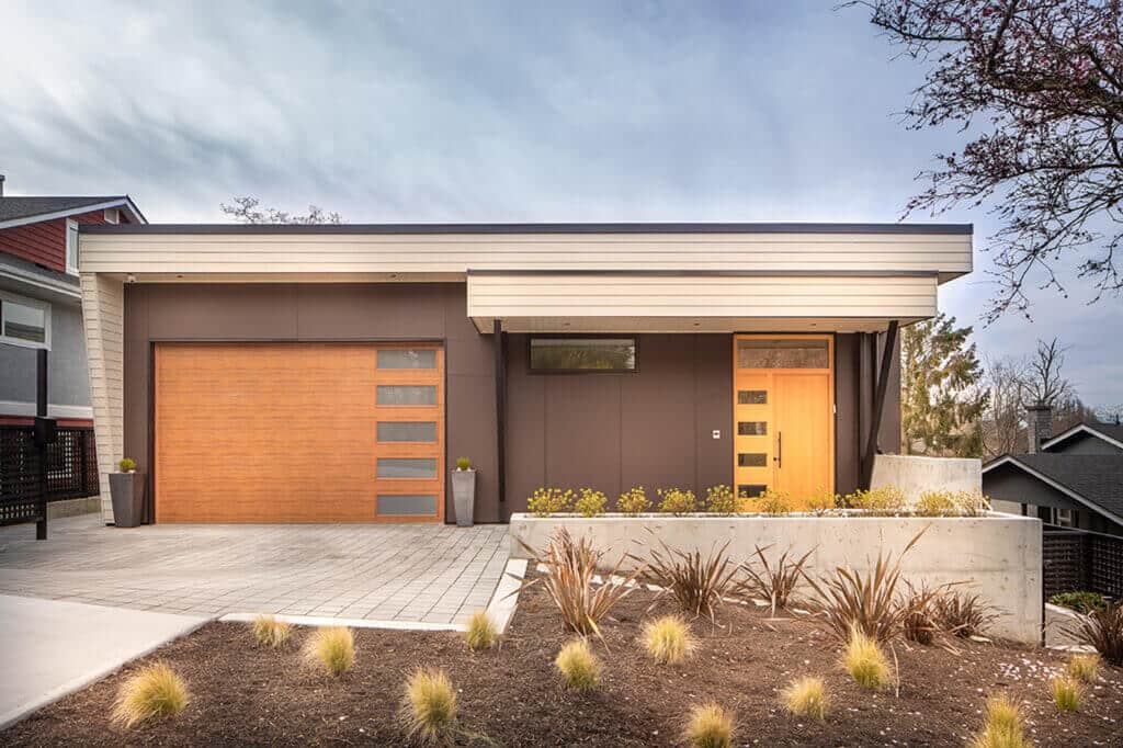 Single-story modern house with flat roof, light brown garage door, matching front door, narrow horizontal window, small garden, and sloped driveway in front.