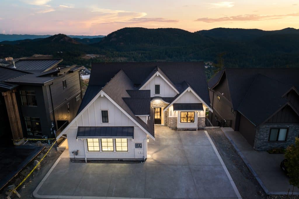 Aerial view of a modern white house with black roof, large driveway, and exterior lighting, set against a backdrop of hills at sunset.