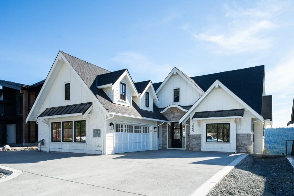 Modern two-story house with white siding, black roof, stone accents, and a three-car garage, set on a large concrete driveway under a clear sky.