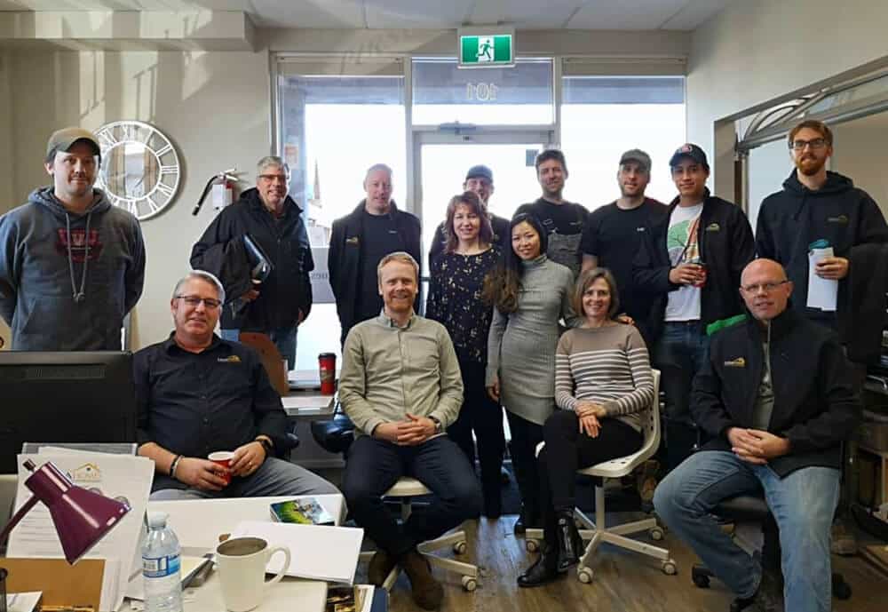 A group of thirteen people, some seated and some standing, gather in an office space with desks, a clock, and a door in the background.