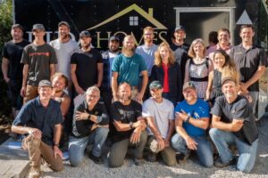 A group of twenty people pose and smile outdoors in front of a trailer with a house logo on it, standing and kneeling on gravel.