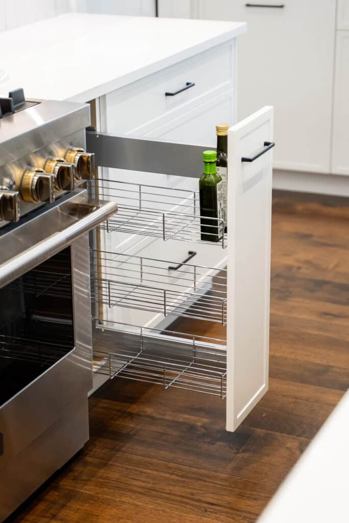 A kitchen cabinet with a pull-out wire rack is partially open, holding two bottles next to a stainless steel oven on a wooden floor.