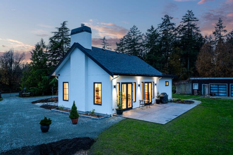 Modern custom home exterior view at dusk, showcasing a stylish design with large windows, a well-maintained yard, and surrounding trees. The inviting patio area features outdoor seating, enhancing the home's appeal.