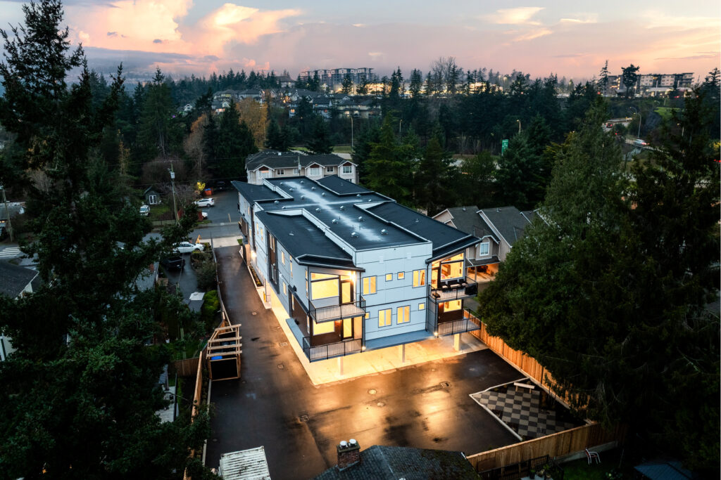 Aerial view of a modern Custom Home featuring a sleek design, surrounded by trees and a residential neighborhood, highlighting its well-lit exterior and spacious parking area.
