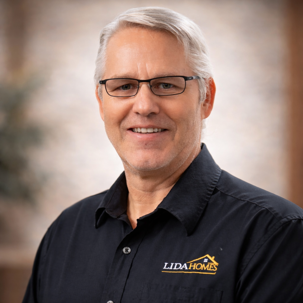 Portrait of a professional man wearing glasses and a black shirt with a logo, representing a Custom Home builder. The background features a softly blurred indoor setting, highlighting his expertise in Custom Home design and construction.